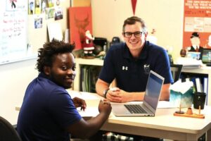 Two men sit at a desk in an office, with a laptop open in front of them. Both are smiling and facing the camera. The office has various decorations and papers on the walls.