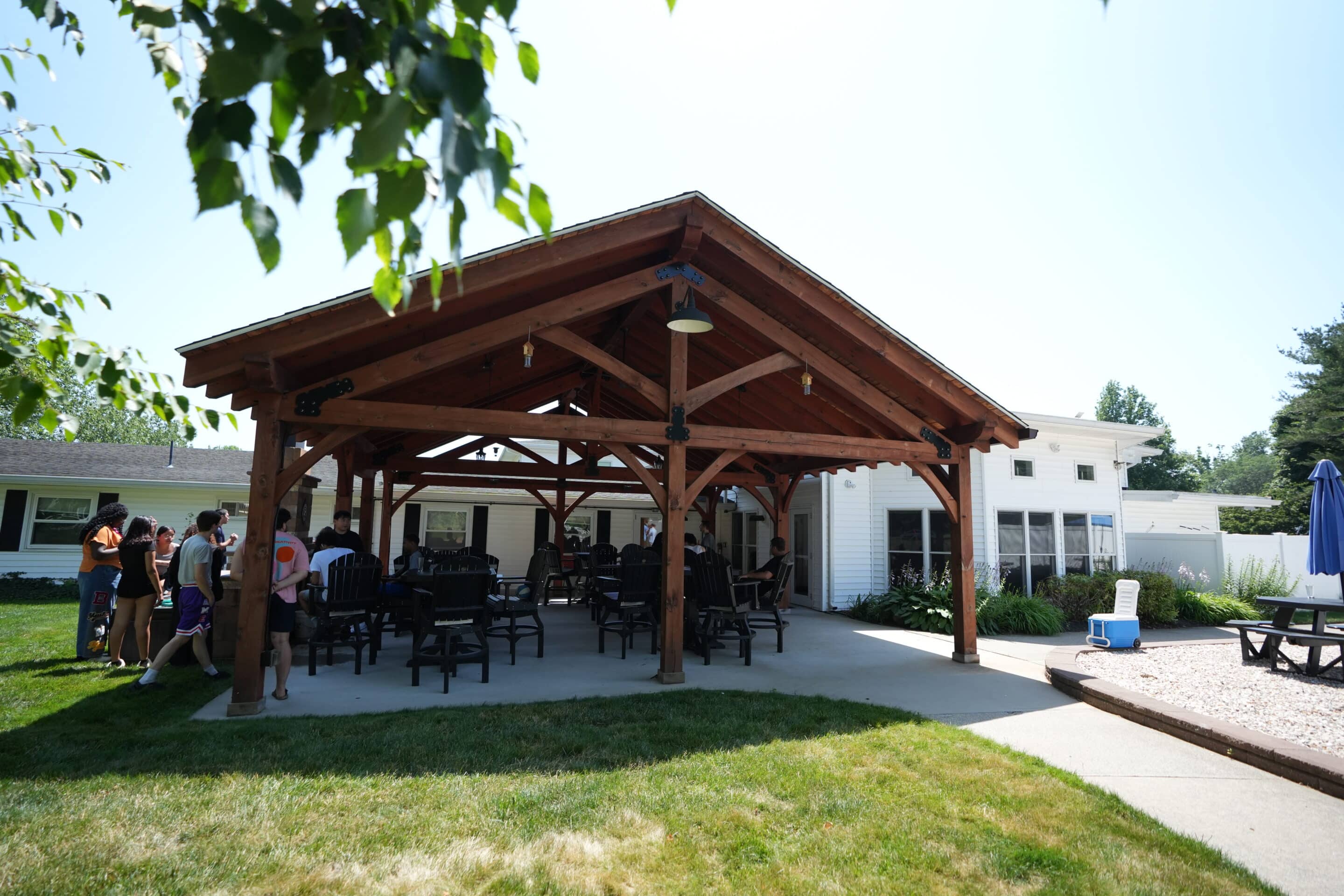 A group of people gathers under a wooden pavilion with tables and chairs beside a white building on a sunny day.