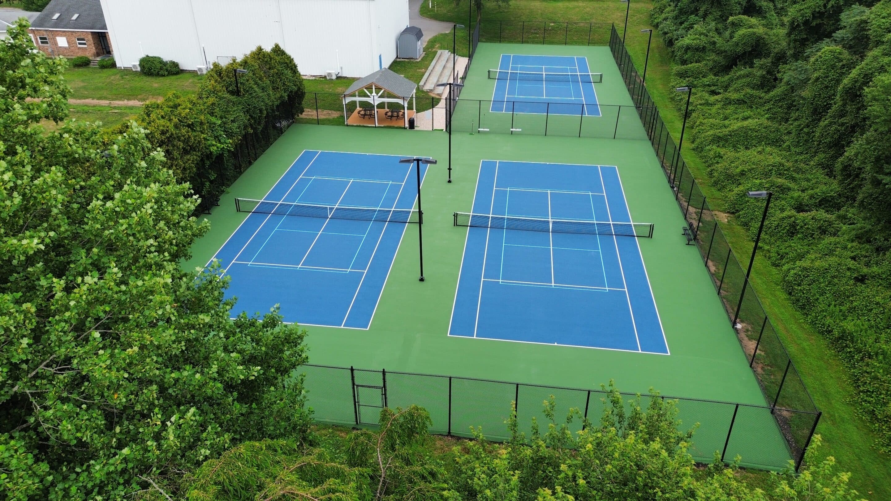 Aerial view of two blue and green outdoor tennis courts surrounded by trees and a fence, with a small shed nearby.