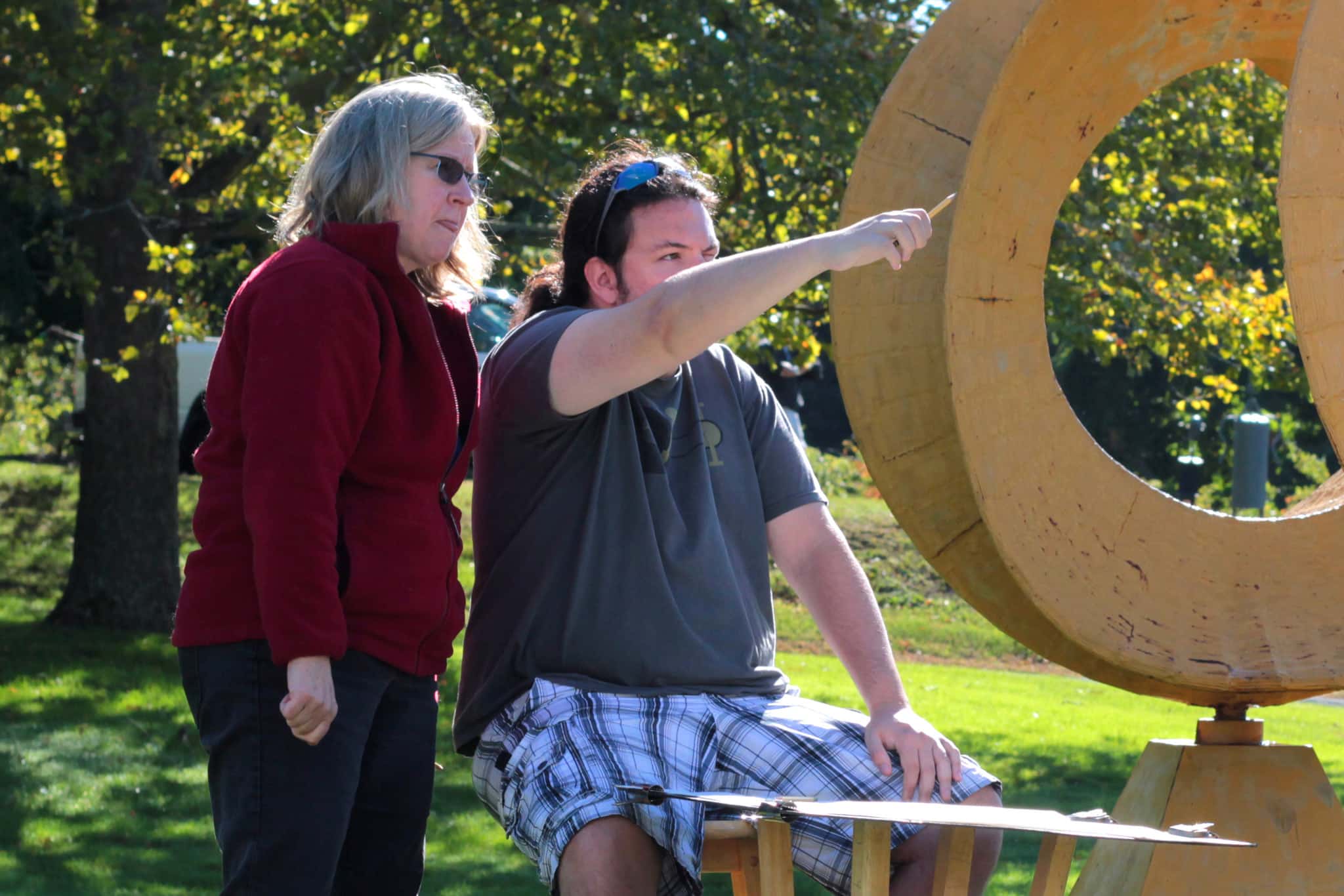 A man sits and points while a woman stands beside him outdoors near a large wooden sculpture on a sunny summer day at Oxford Academy.